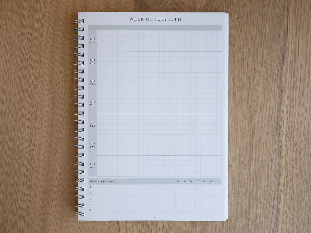 A spiral-bound weekly planner page from a larger white planner, laid flat on a light brown wood-grain surface. The page is headed 'WEEK OF JULY 13TH' and features a grid layout for tracking daily habits and tasks.