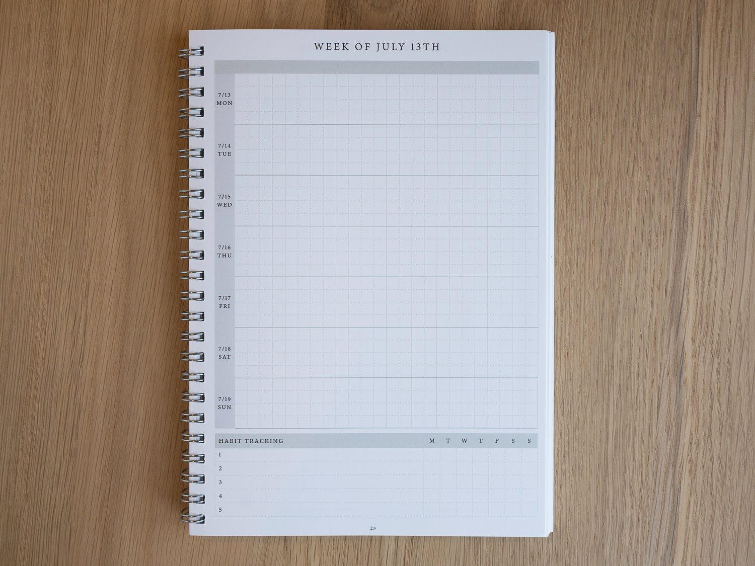 A spiral-bound weekly planner page from a larger white planner, laid flat on a light brown wood-grain surface. The page is headed 'WEEK OF JULY 13TH' and features a grid layout for tracking daily habits and tasks.