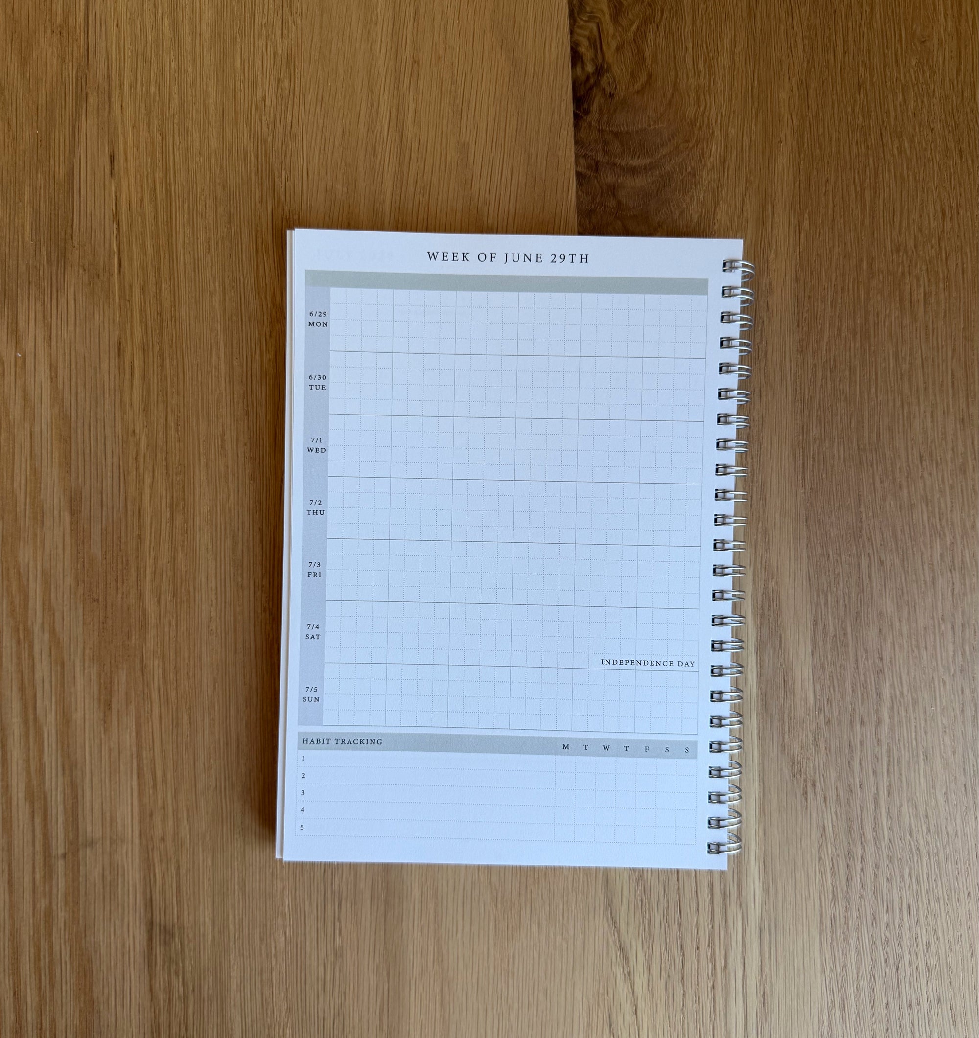 A spiral-bound weekly planner pad with a white page and light gray grid lines, placed on a light brown wooden surface.